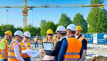 The image depicts a modern construction site bustling with activity under a clear blue sky In the foreground a group of diverse workers in hard hats and safety vests is gathered around a digital tablet reviewing project plans Behind them towering cra-3