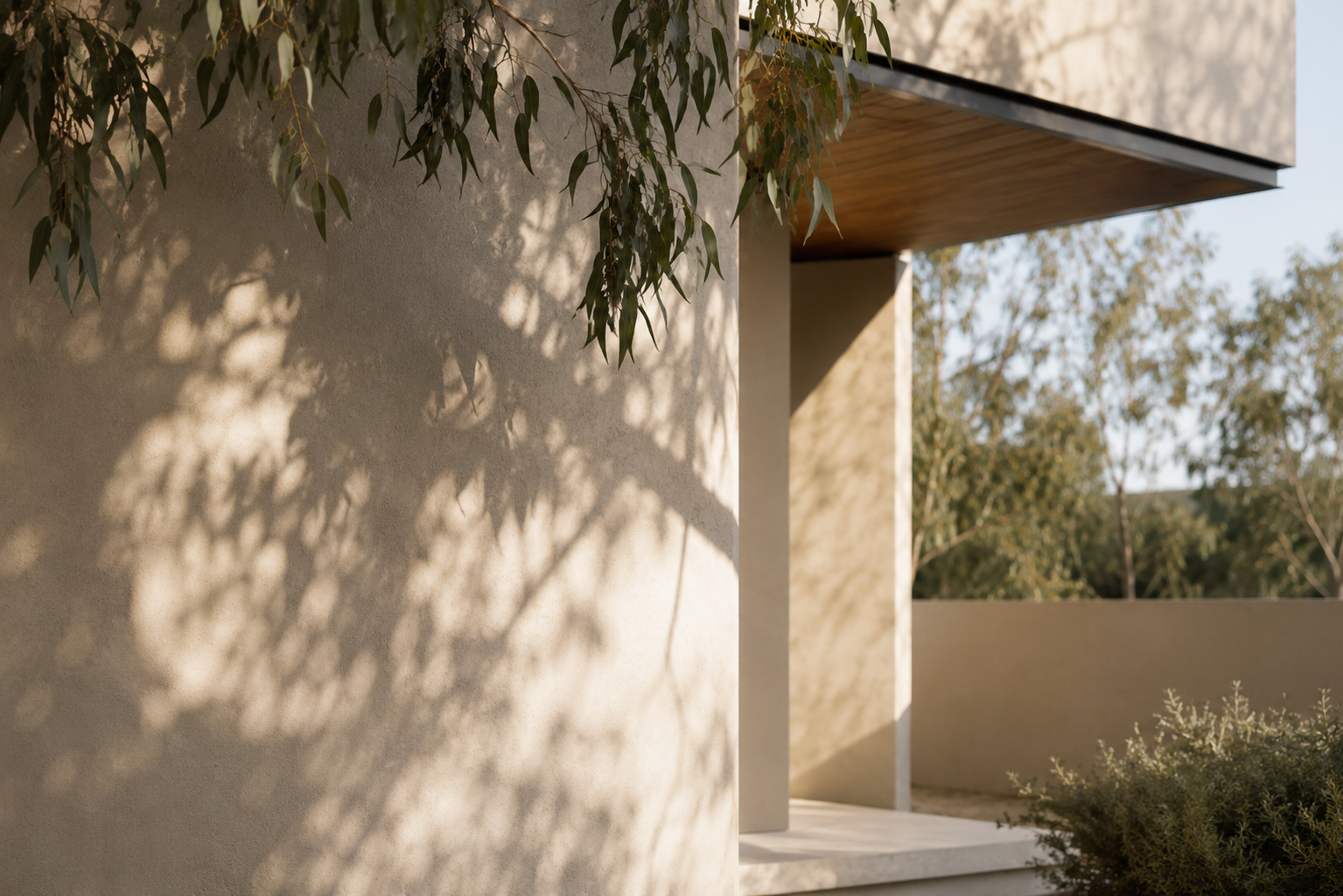 Residential façade detail with rendered walls, deep roof overhang and filtered tree shading for solar control.